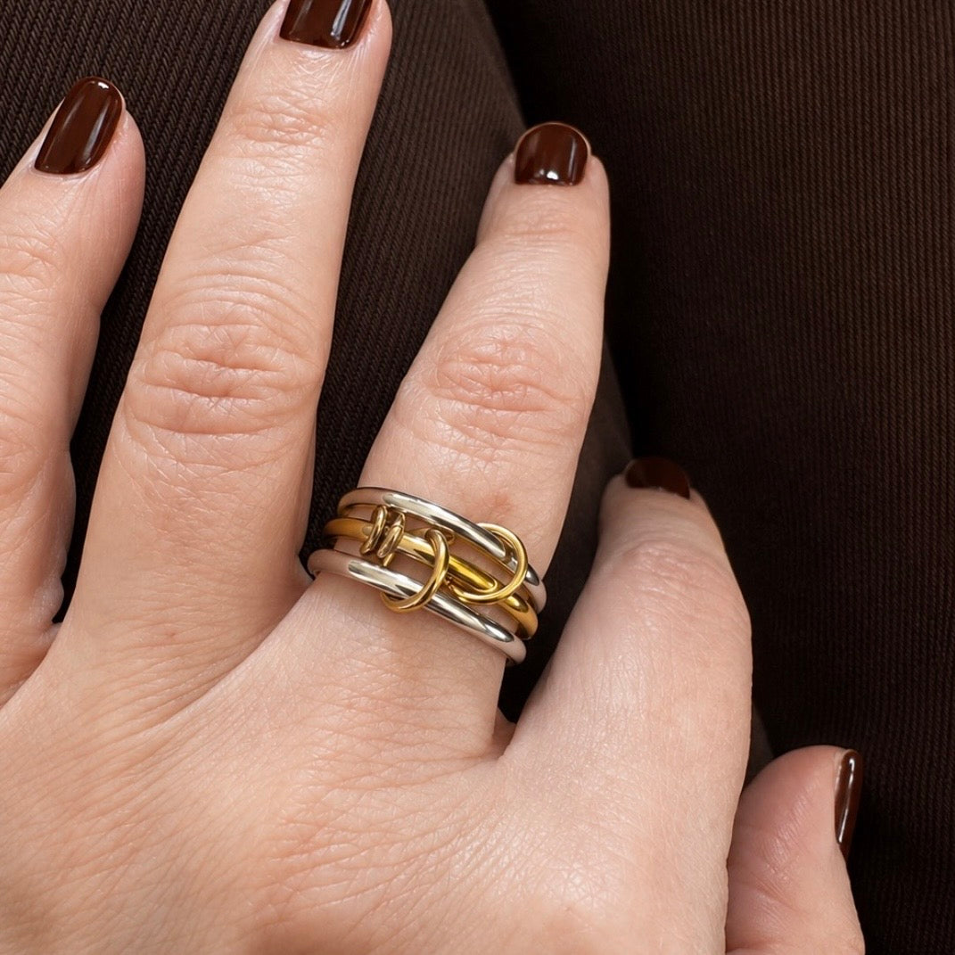 A close-up of a hand wearing a multi-band ring with interlocking gold and silver loops, resting on dark brown clothing.