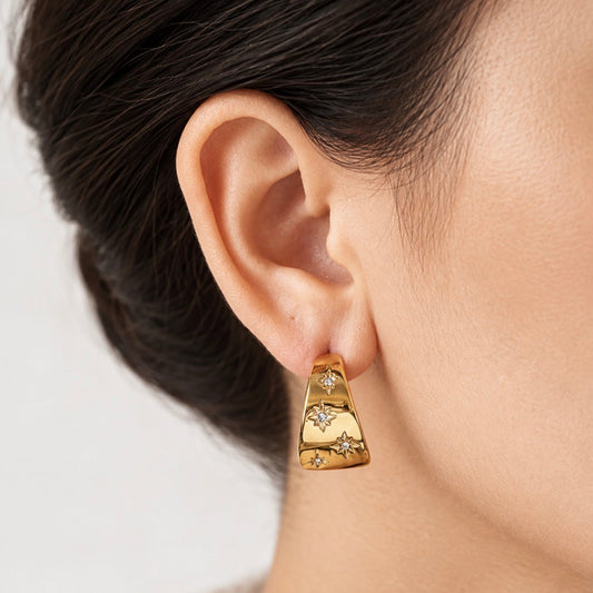 A close-up of a model's ear wearing a gold crescent-shaped earring adorned with small sparkling star and diamond-like accents.