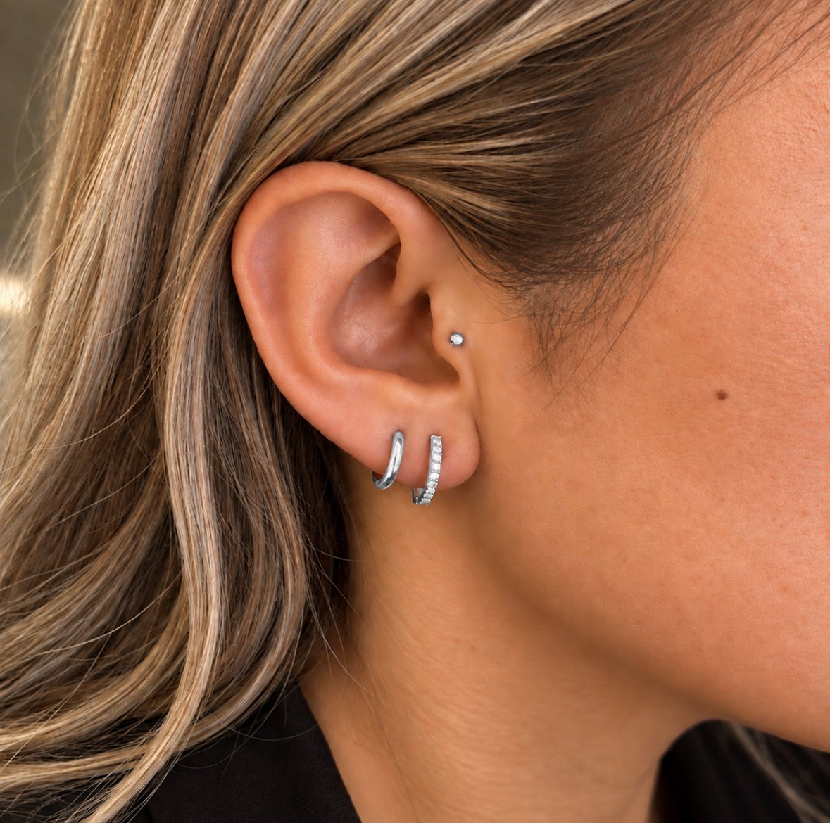 Close-up of a person's ear wearing multiple silver hoop and stud earrings, including a small silver nose stud, with blonde hair and skin visible.