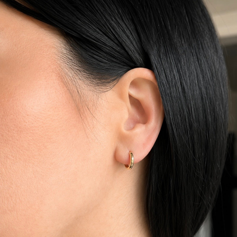 A close-up of a person's ear wearing a small gold huggie hoop earring, with dark hair and smooth skin visible.