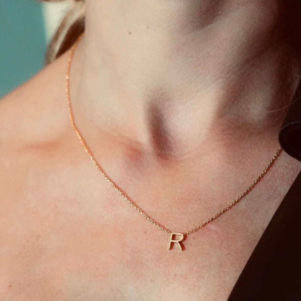 Gold necklace with a 'R' pendant on a person's neck against a neutral background