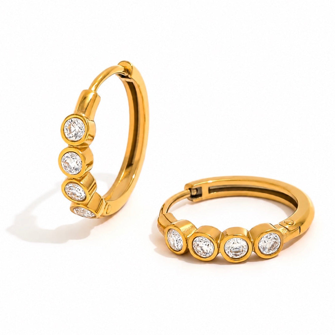 A pair of gold hoop earrings, each adorned with four round-cut clear gemstones in bezel settings, displayed on a clean white background.