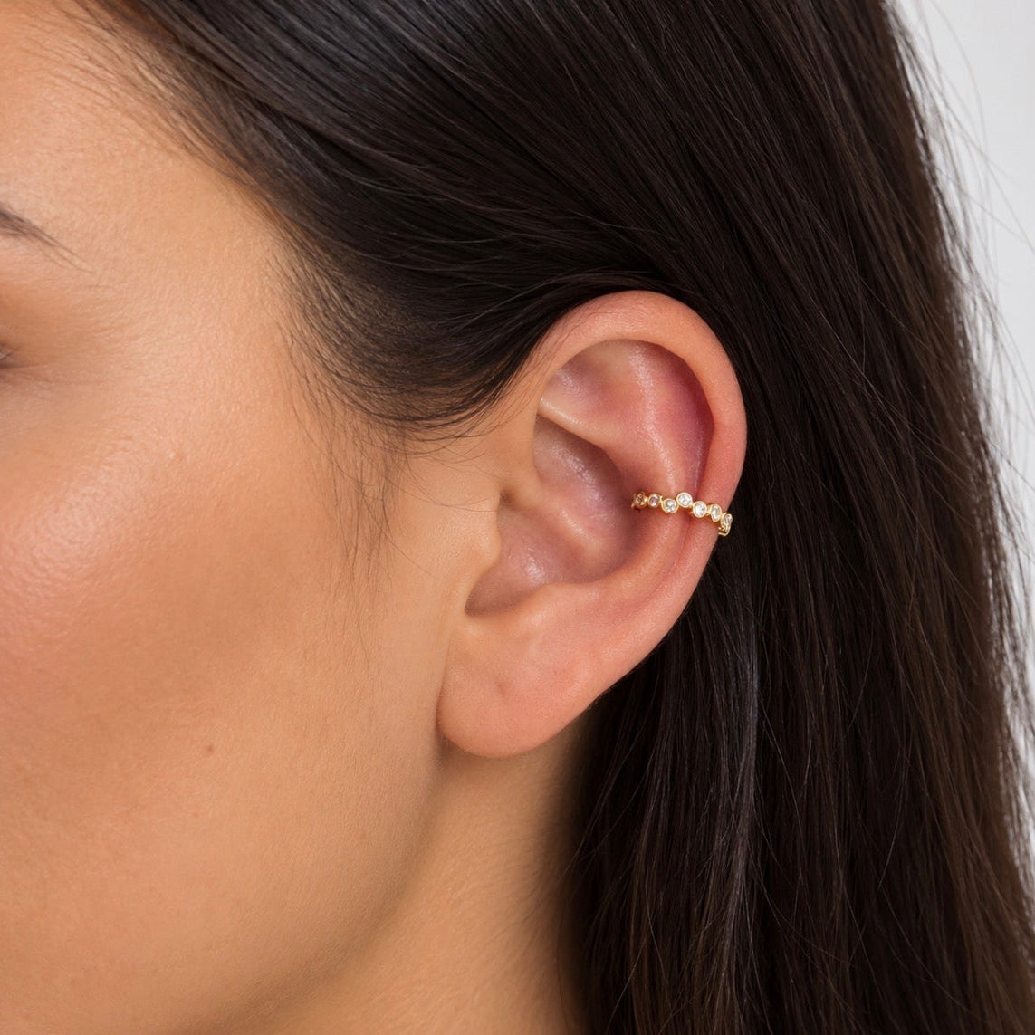 A close-up of a woman's ear wearing a delicate gold ear cuff adorned with small clear stones.