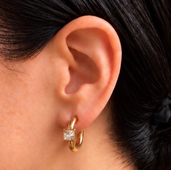 A close-up of a person's ear wearing a gold huggie hoop earring with a square-cut clear gemstone.