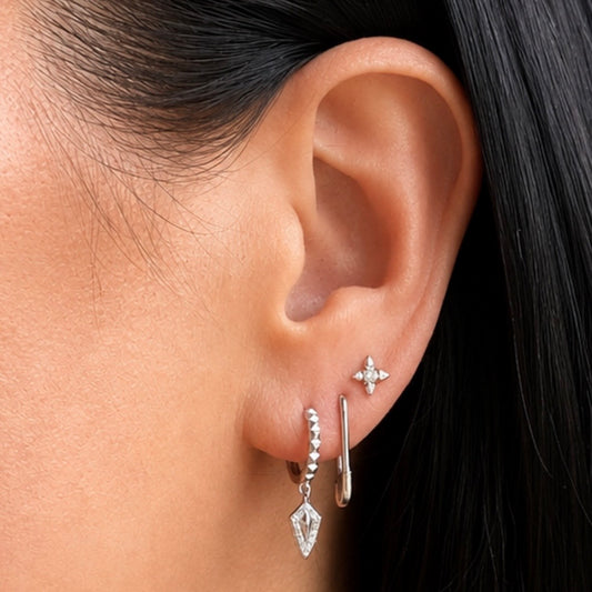 A close-up of a person's ear showcasing silver hoop and stud earrings. The main earring is a silver huggie hoop adorned with tiny clear stones and a dangling clear crystal pendant in a diamond shape.