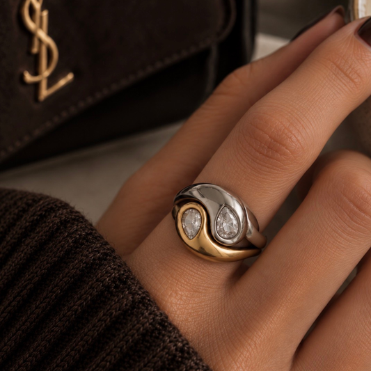 A close-up of a hand wearing a two-tone yin-yang style ring with diamond accents, set against a dark brown textured sweater and a black YSL handbag in the background.