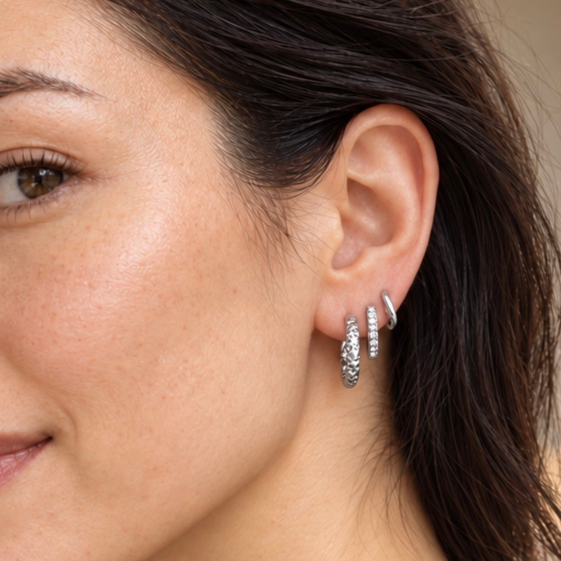 Close-up of a woman's ear wearing three silver hoop earrings, including one textured silver hoop and two smaller silver huggie hoops.
