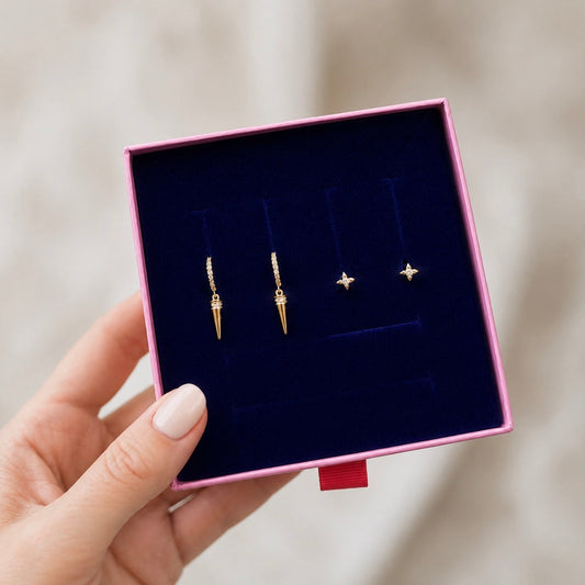 A hand holding an open pink jewelry box with a dark blue velvet insert, displaying three pairs of gold-toned earrings against a soft beige background.