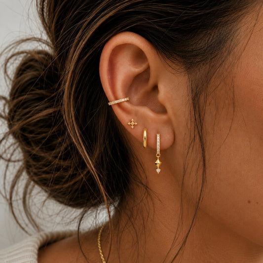 A close-up of a woman's ear wearing multiple delicate gold earrings, including a small gold hoop, a dangling gold charm with a tiny clear stone, a thin horizontal clear-stone ear cuff, and a small star-shaped stud.