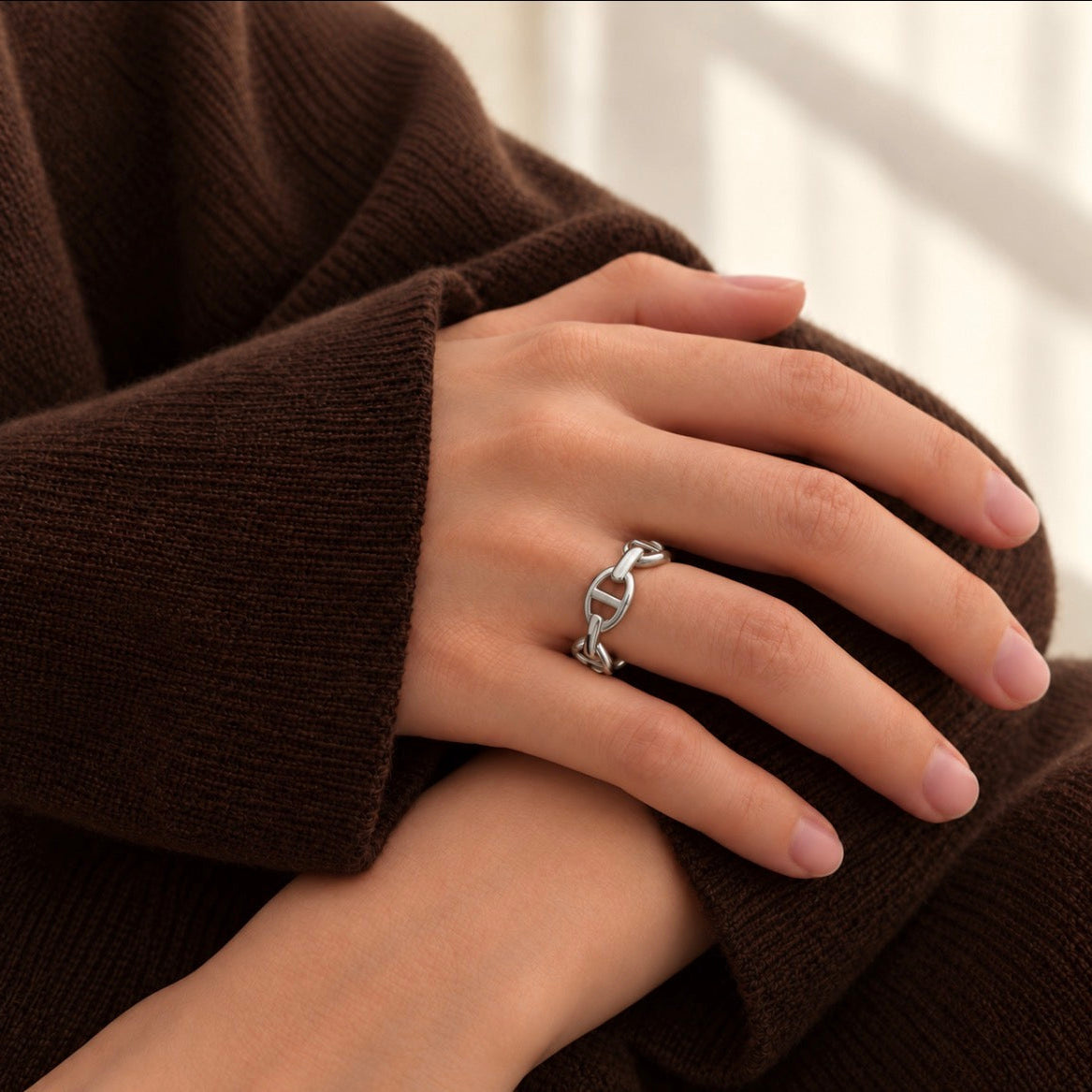 A close-up of a person's hand wearing a silver ring with a interlocking anchor link design, resting on a brown ribbed knit sweater.