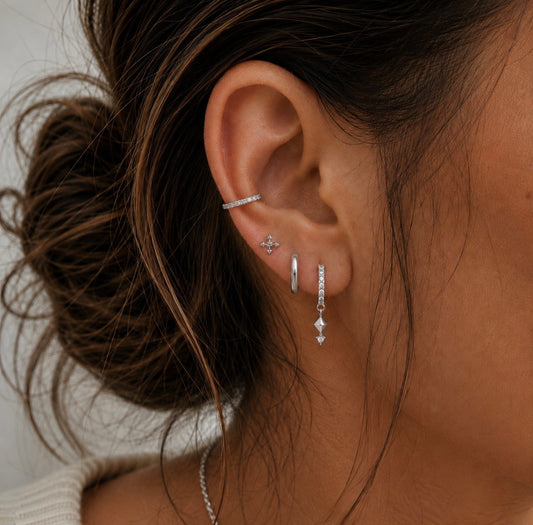 A close-up of a person's ear wearing multiple delicate silver earrings, including a small diamond stud, a huggie hoop, a barbell cartilage piercing, and a long dangling huggie with a diamond-accented drop.
