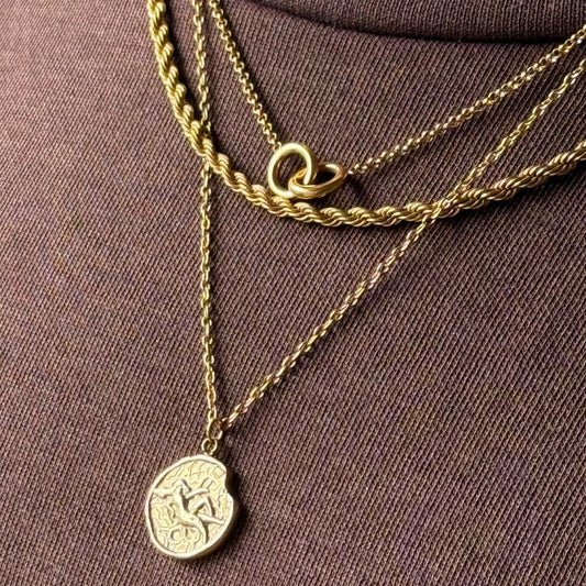 Gold necklaces with a zodiac coin pendant layered with rope chain necklace and double ring necklace on a model brown fabric background