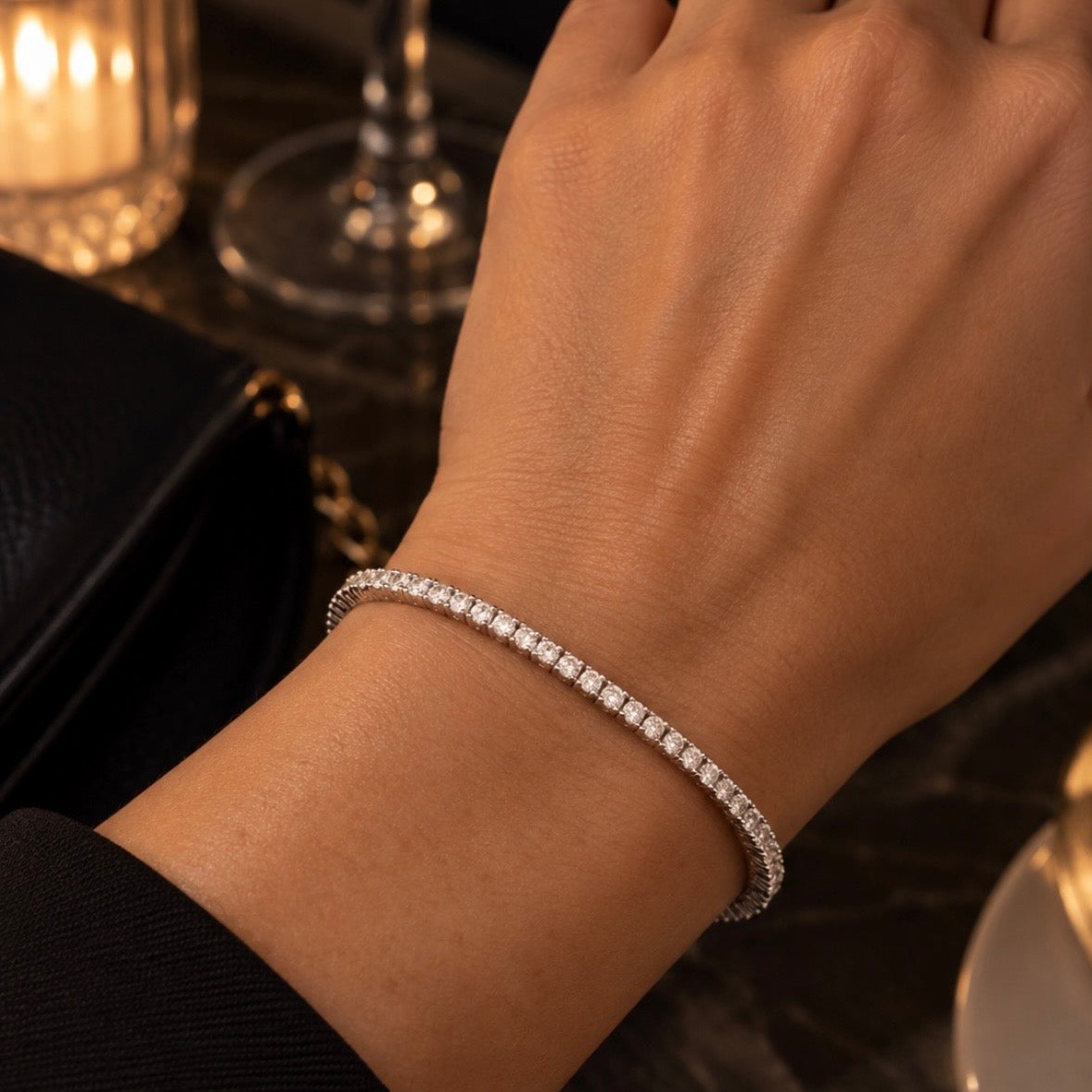 A close-up of a person's wrist wearing a thin, diamond-encrusted tennis bracelet in a warm, dimly lit setting.
