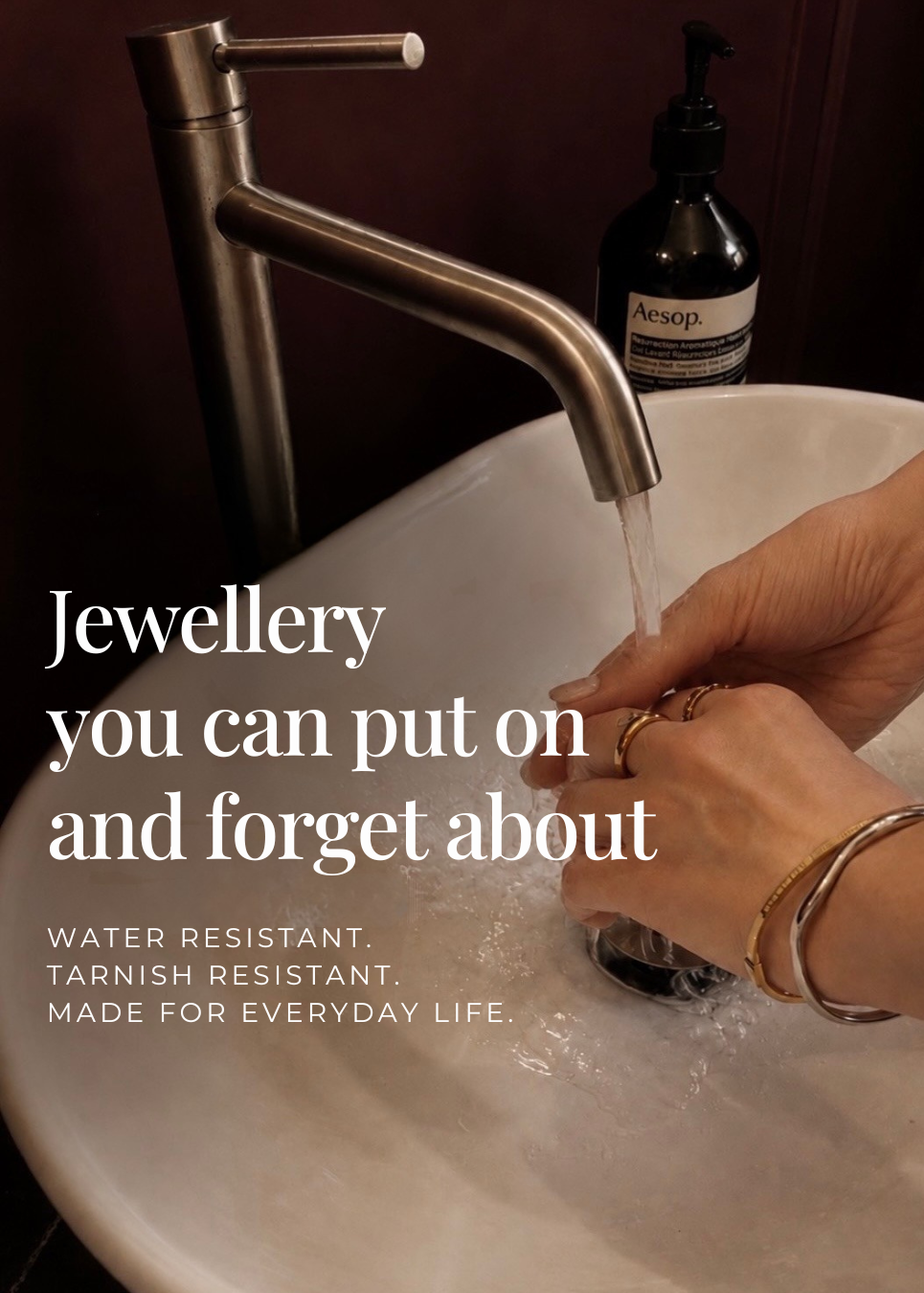 A person's hands under running water from a modern brushed nickel faucet in a white sink, wearing gold bangles and rings. A dark bottle of Acneff soap is visible in the background.