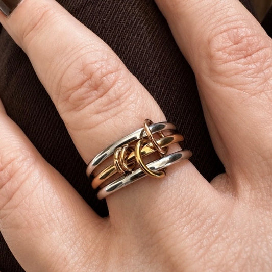 A close-up of a hand wearing a multi-band ring with interlocking silver and gold-toned bands, set against a dark brown textured background.