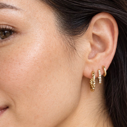 Close-up of a model's ear wearing three gold hoop earrings, including one textured gold hoop, one small gold hoop with clear stones, and a gold ear cuff.