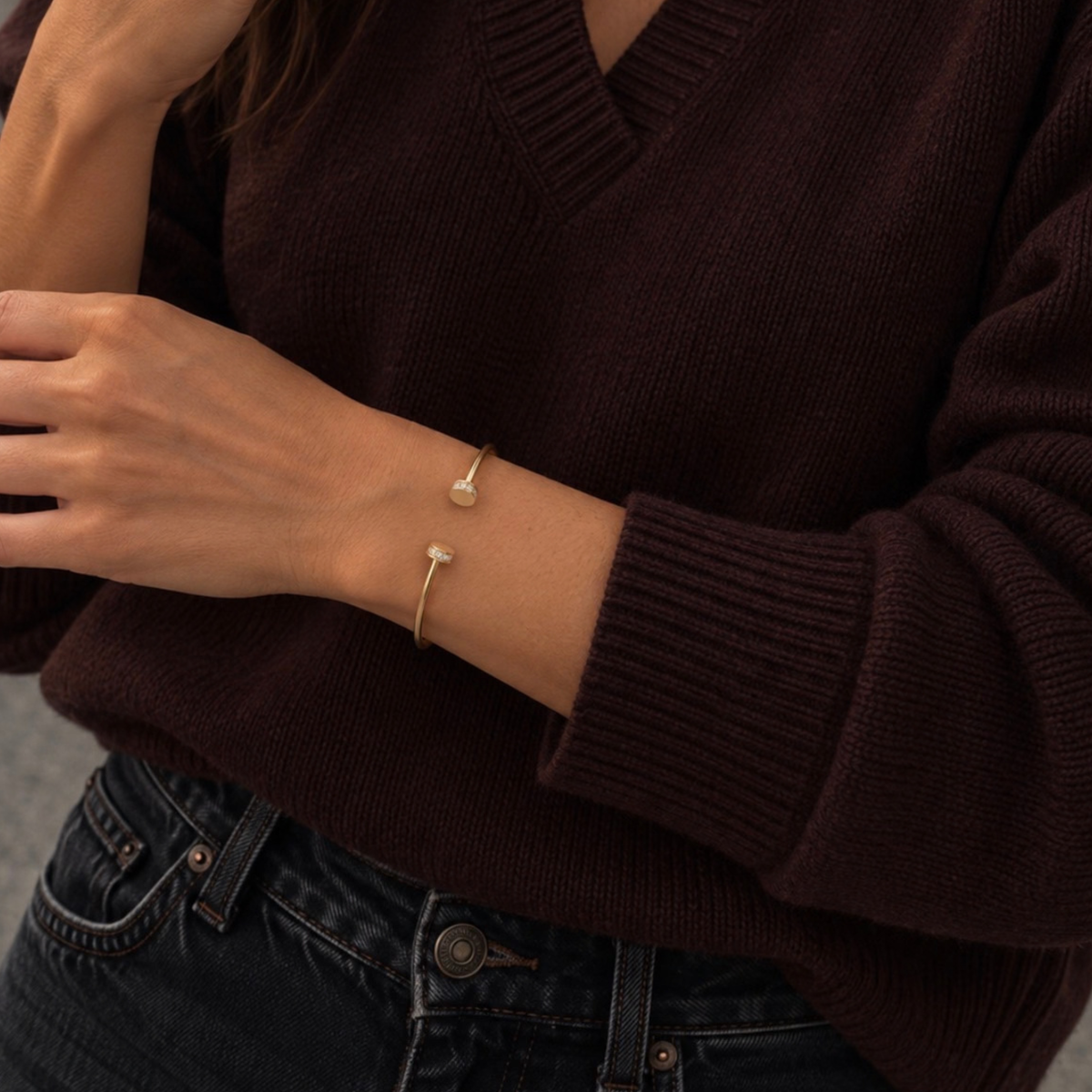 A close-up of a person wearing a delicate gold bangle bracelet with a small pearl and a tiny gold star, paired with a dark brown ribbed knit sweater and dark blue jeans.