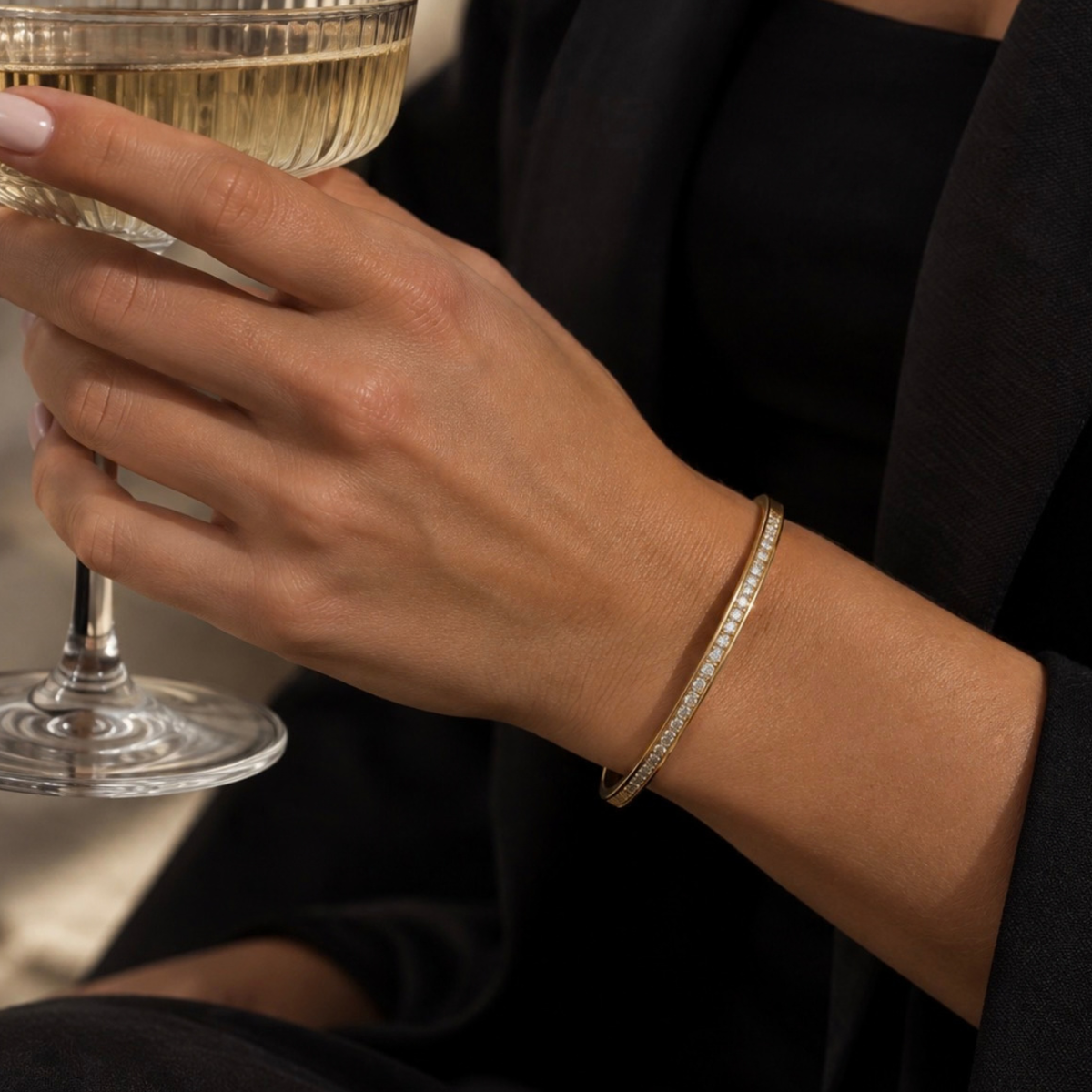 A close-up of a woman's hand holding a glass of white wine while wearing a delicate gold bangle with small clear stones on her wrist.