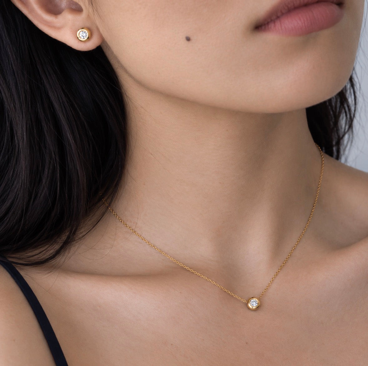 A close-up lifestyle shot of a model wearing a delicate gold chain necklace with a single round-cut clear gemstone pendant, along with a matching gold stud earring.