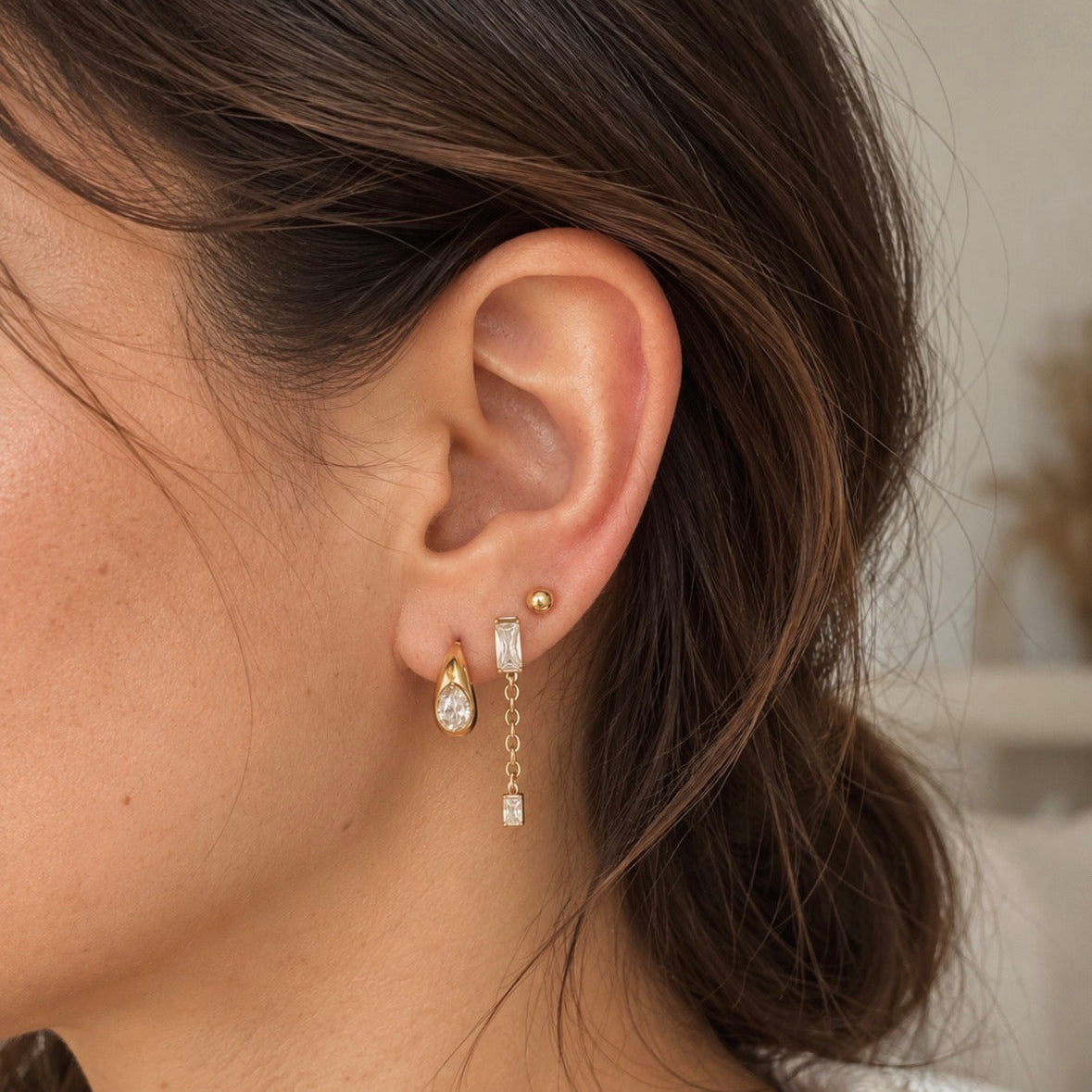 A close-up of a woman's ear wearing a delicate gold dangle earring with clear teardrop-shaped stones and a small gold stud, against a softly blurred indoor background.