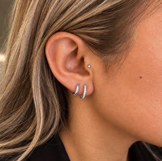Close-up of a person's ear wearing multiple silver hoop and stud earrings, including a small silver nose stud, with blonde hair and skin visible.