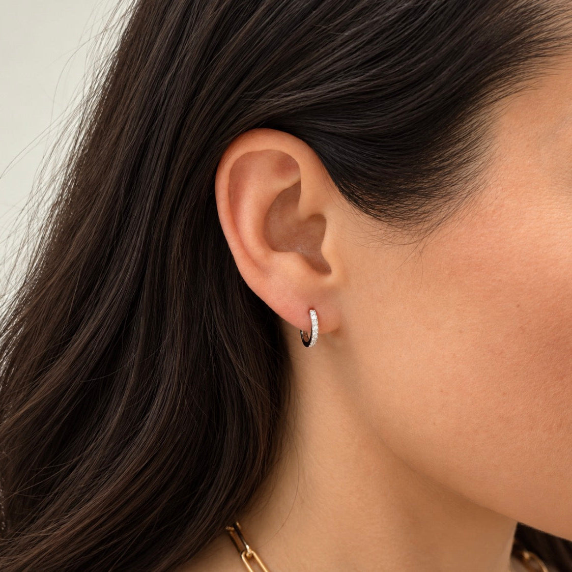 A close-up of a model's ear wearing a small silver huggie hoop earring with a pavé setting, shown against dark brown hair and light skin.