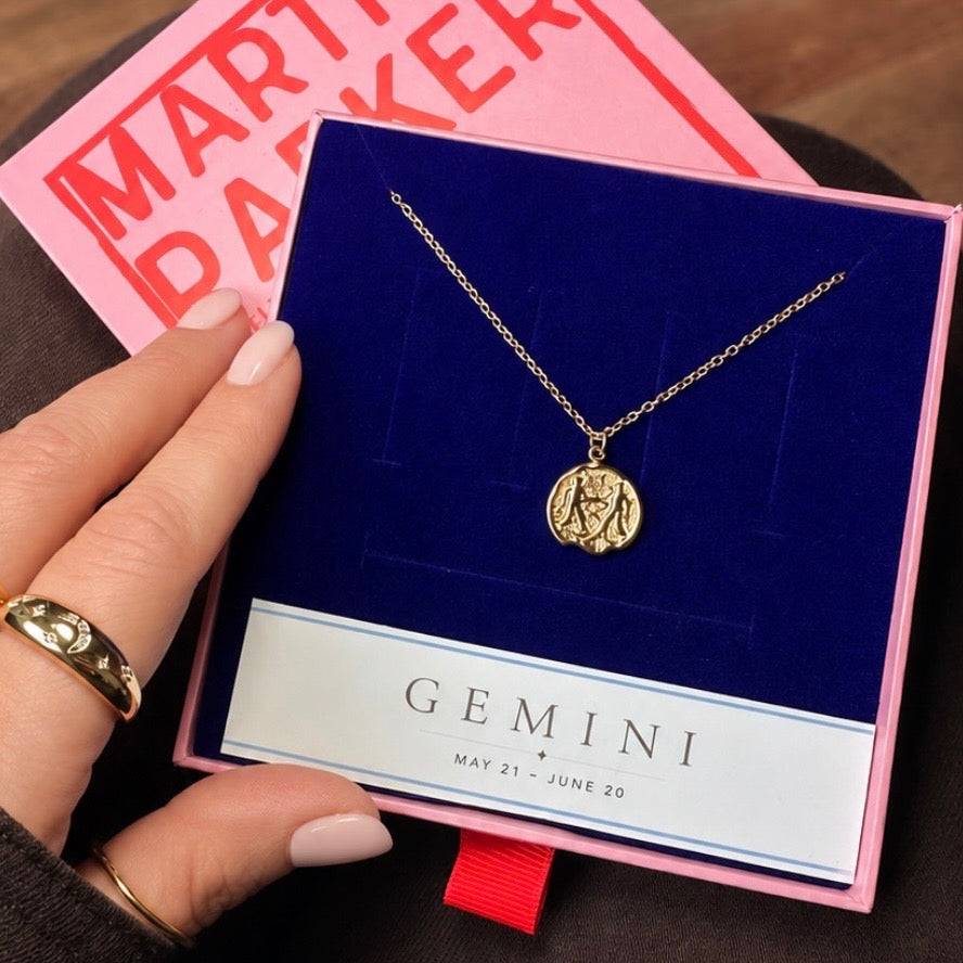 Gold necklace with gemini pendant in branded Martha Parker gift box on a blue card held by a hand, with a pink box in the background.