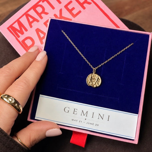 Gold necklace with gemini pendant in branded Martha Parker gift box on a blue card held by a hand, with a pink box in the background.