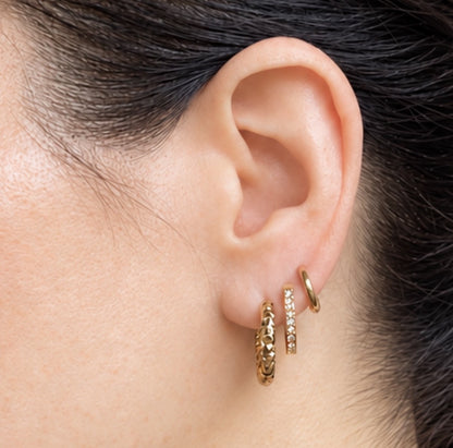 A close-up of a person's ear wearing three gold hoop earrings, including one textured gold hoop, one gold hoop with small clear stones, and one thin gold hoop.