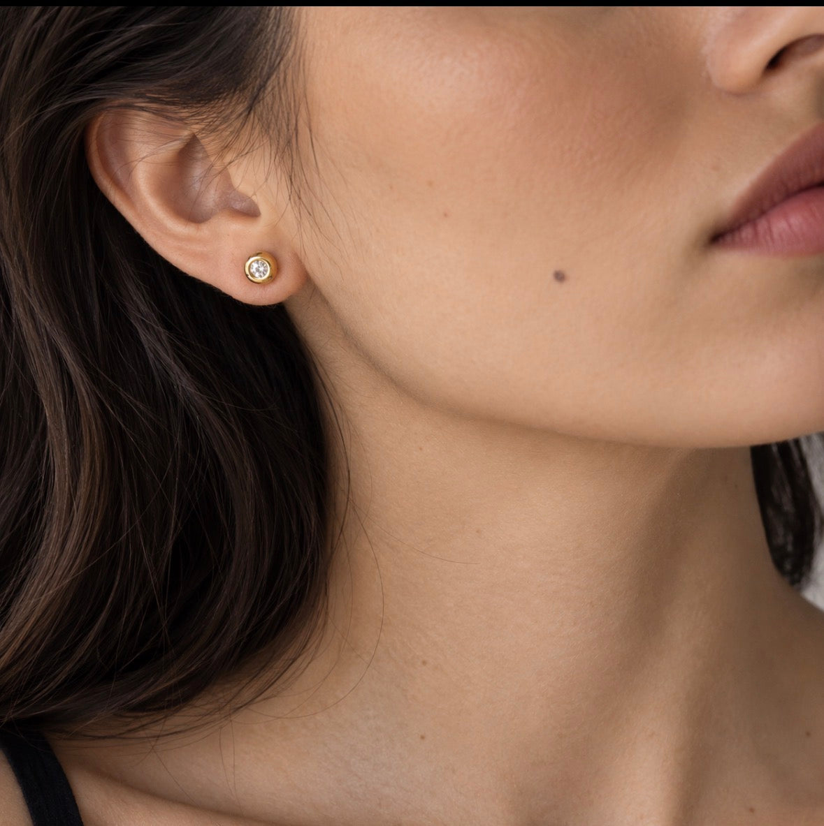 A close-up of a woman's ear wearing a small gold stud earring with a clear gemstone, shown against her skin and dark hair.
