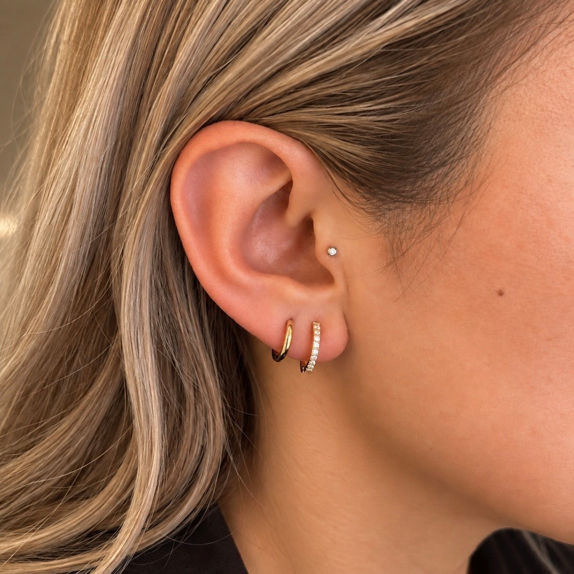 Close-up of a person's ear wearing two small hoop earrings, one plain gold and one with tiny clear stones, plus a small stud earring on the upper ear.
