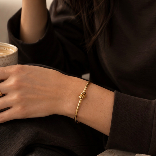 A delicate gold bangle bracelet with a knotted center detail, worn on a person's wrist. The person is wearing a dark brown or black long-sleeve top and holding a ceramic cup, captured in a warm, cozy indoor setting.