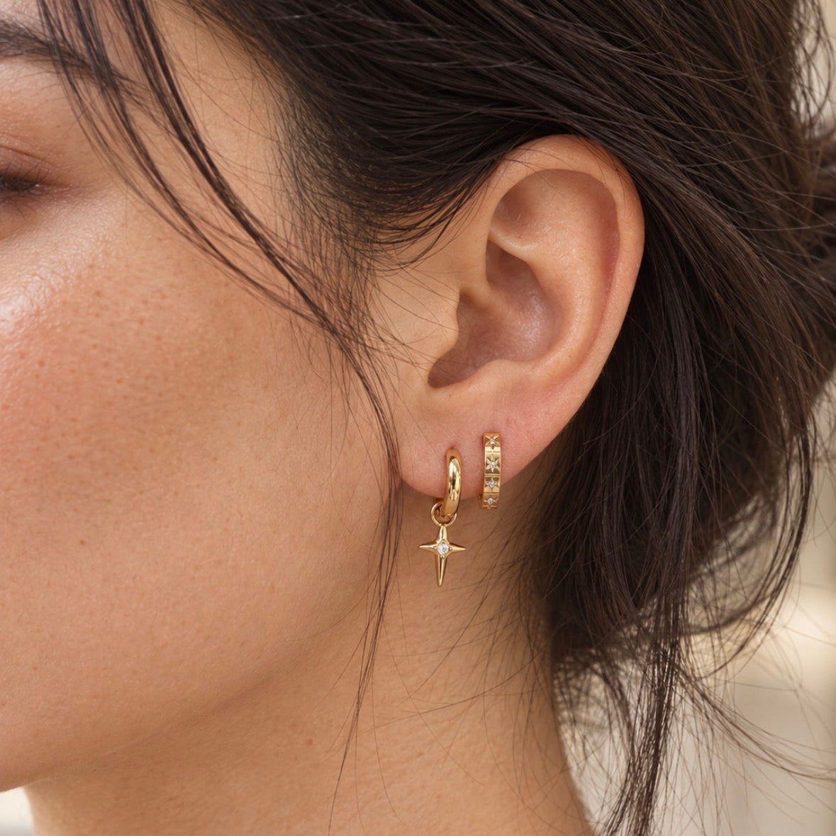 A close-up side profile of a woman wearing a pair of gold hoop earrings, one of which features a small dangling star charm and the other a small gold ring set with tiny clear stones.