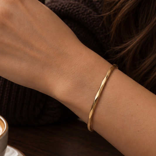A thin twisted gold bangle bracelet worn on a person's wrist, shown in a close-up lifestyle shot with a dark knit sweater and a coffee cup in the background.