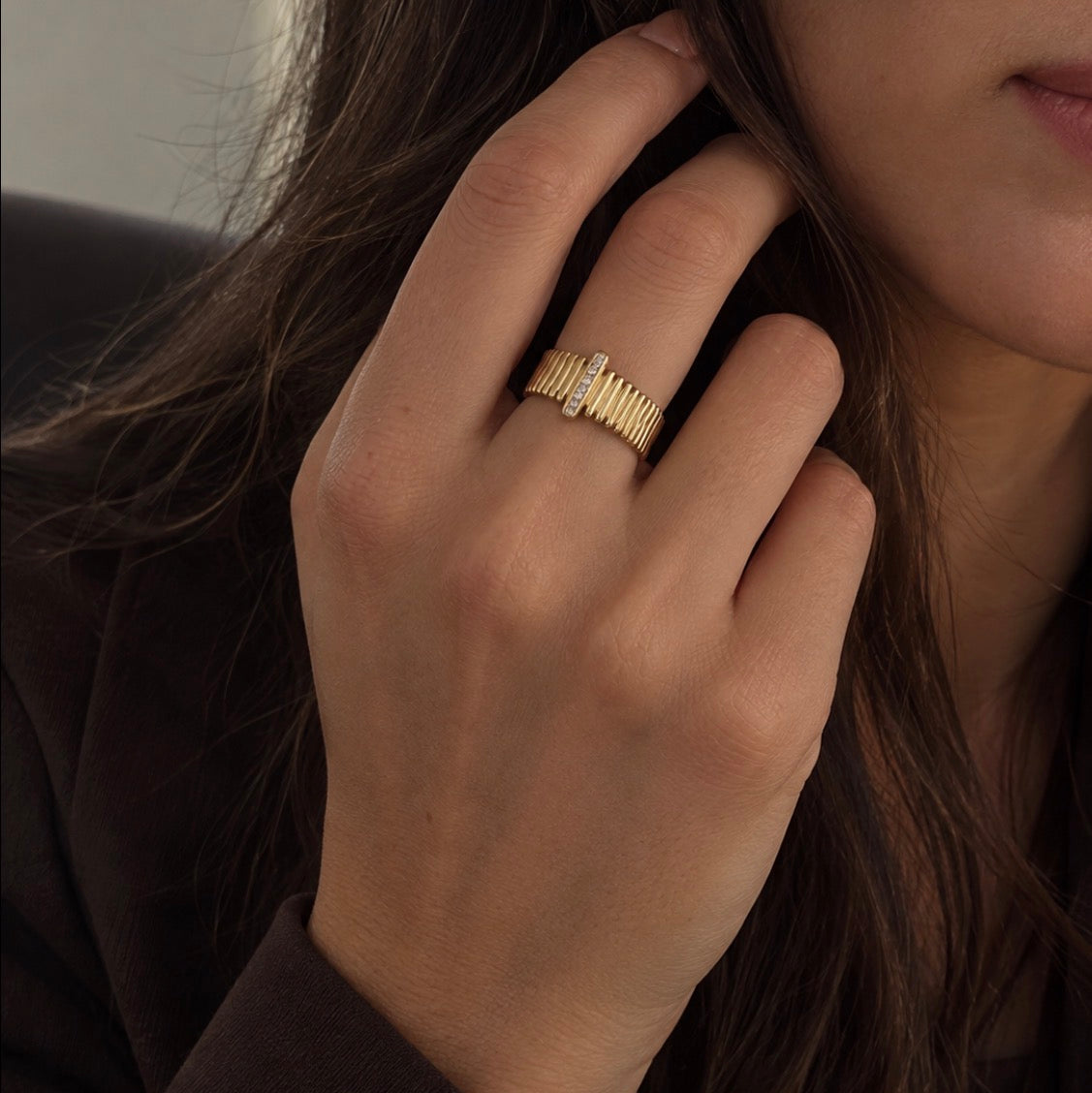 A close-up of a person's hand wearing a textured gold ring with small clear stones, set against dark brown hair and a dark brown jacket.