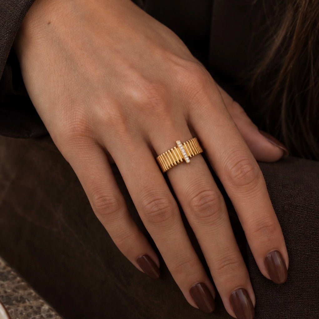 A close-up of a person's hand wearing a gold ring with a textured, segmented design and a small central clear stone, set against a dark brown background.