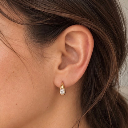 A close-up of a model's ear wearing a small gold teardrop stud earring with a clear teardrop gemstone.