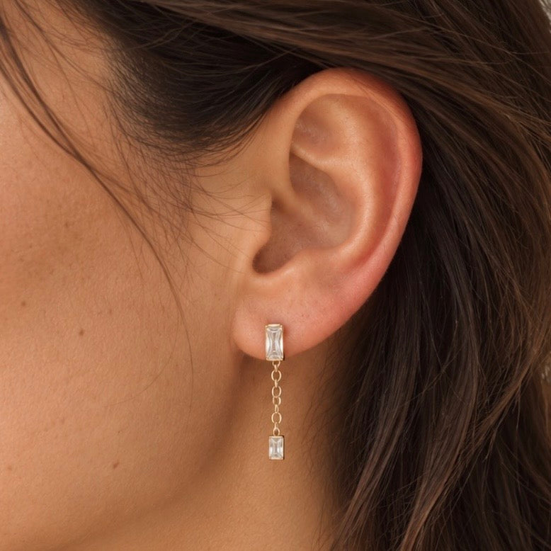 A close-up of a person's ear wearing a gold dangle earring with two clear rectangular baguette gemstones connected by a delicate chain.