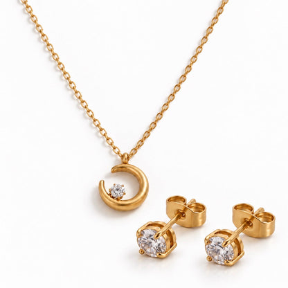 Gold necklace and matching stud earrings set on a white background. The necklace features a crescent moon pendant with a small clear gemstone, and the earrings have round clear gemstones in gold settings.