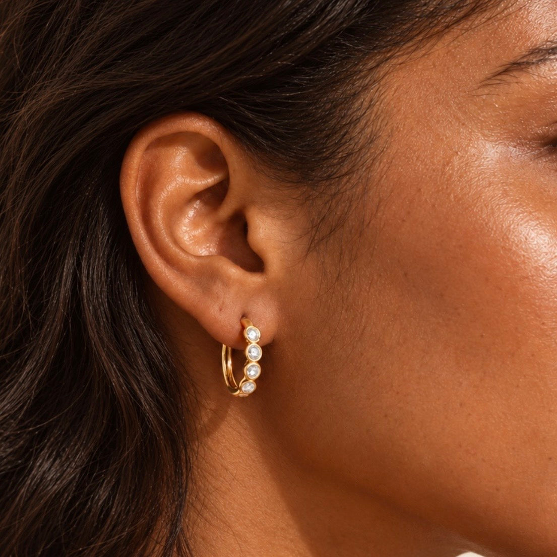 model wearing a gold hoop earring adorned with four round-cut clear stones in bezel settings.