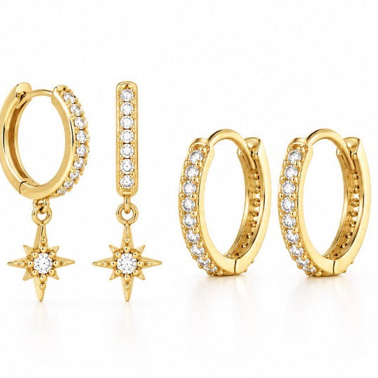 A set of gold huggie hoop earrings embellished with clear pavé stones, including one hoop with a dangling starburst charm and a matching vertical bar with a star charm, displayed on a clean white background.