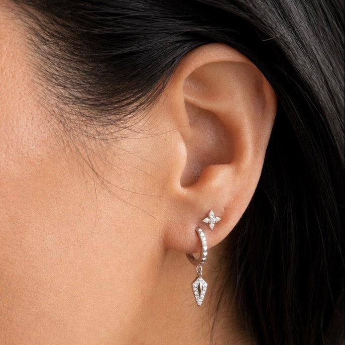 A close-up of a model's ear wearing a silver huggie hoop earring with a dangling chevron-shaped charm and a small starburst stud.