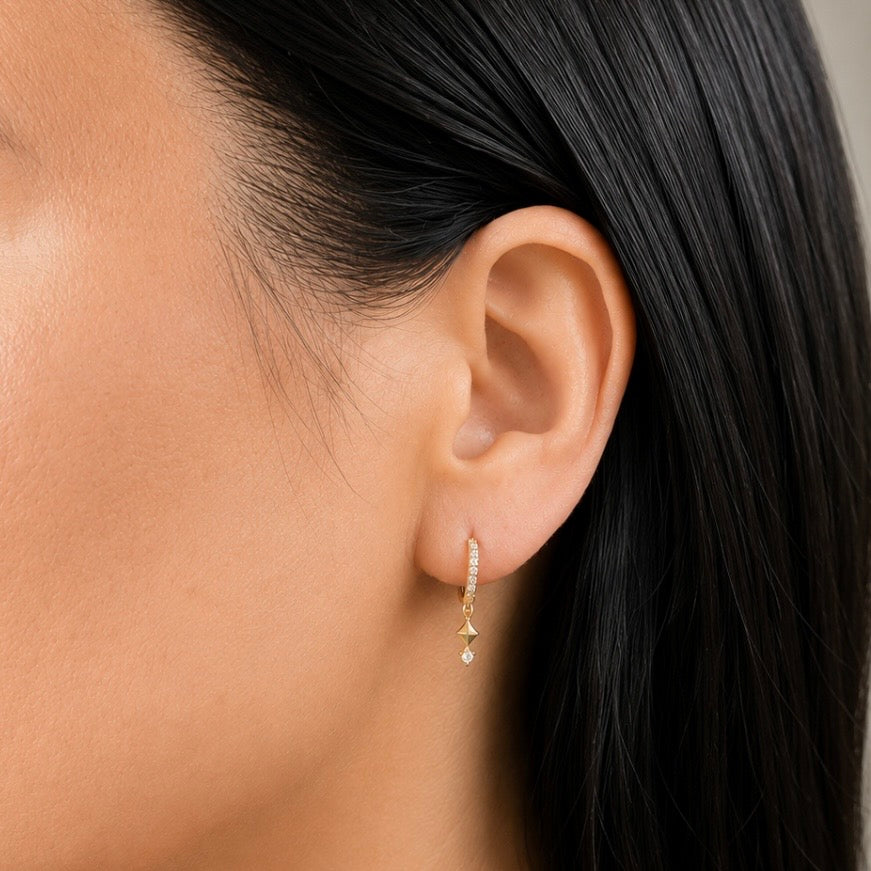 A close-up of a model's ear wearing a delicate gold huggie hoop earring adorned with small clear stones and a dangling gold starburst with a tiny clear teardrop.
