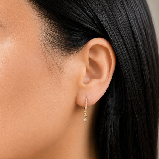 A close-up of a model's ear wearing a delicate gold huggie hoop earring adorned with small clear stones and a dangling gold starburst with a tiny clear teardrop.