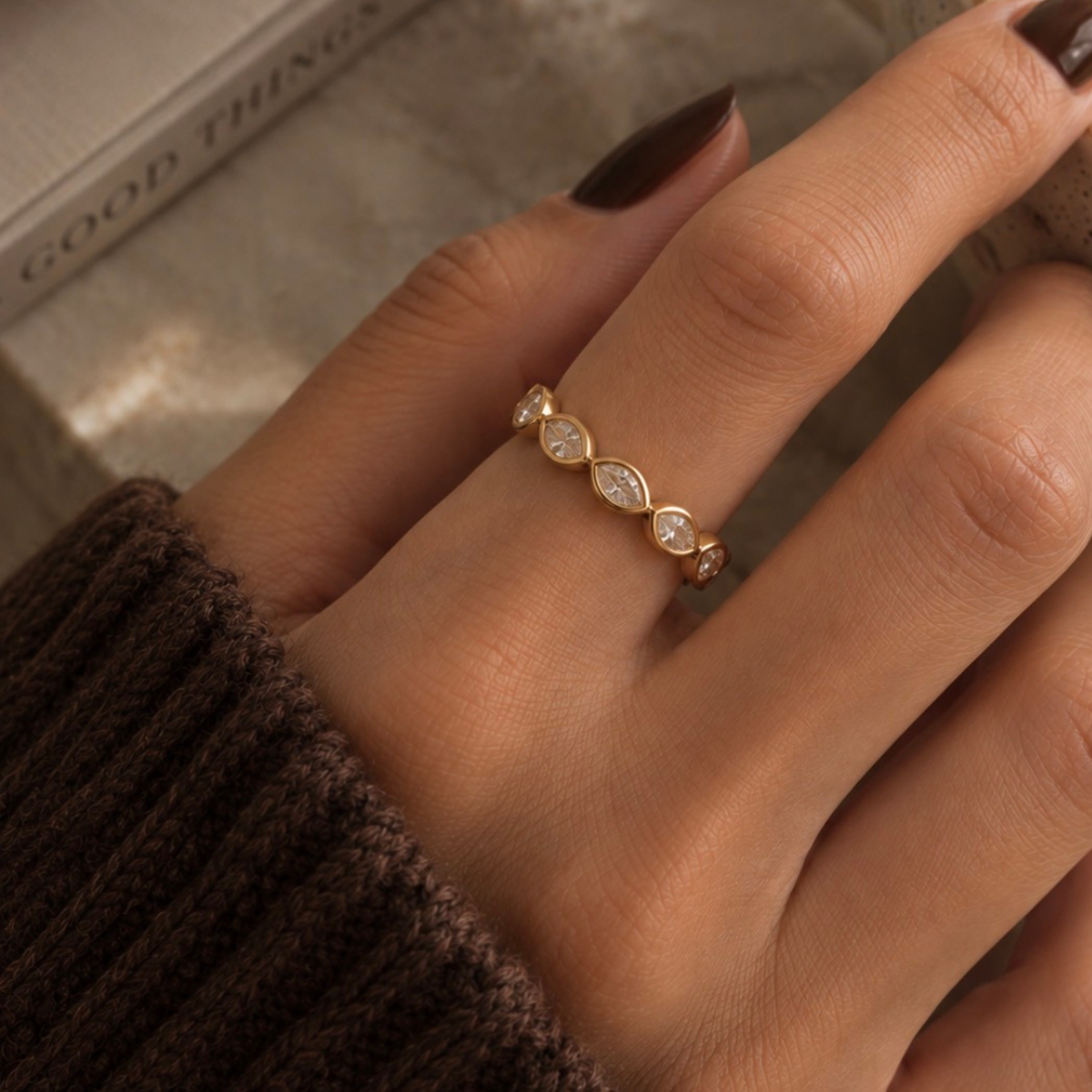 A close-up of a hand wearing a delicate gold ring with three marquise-cut clear stones, styled with a dark brown knit sweater and a book in the background.