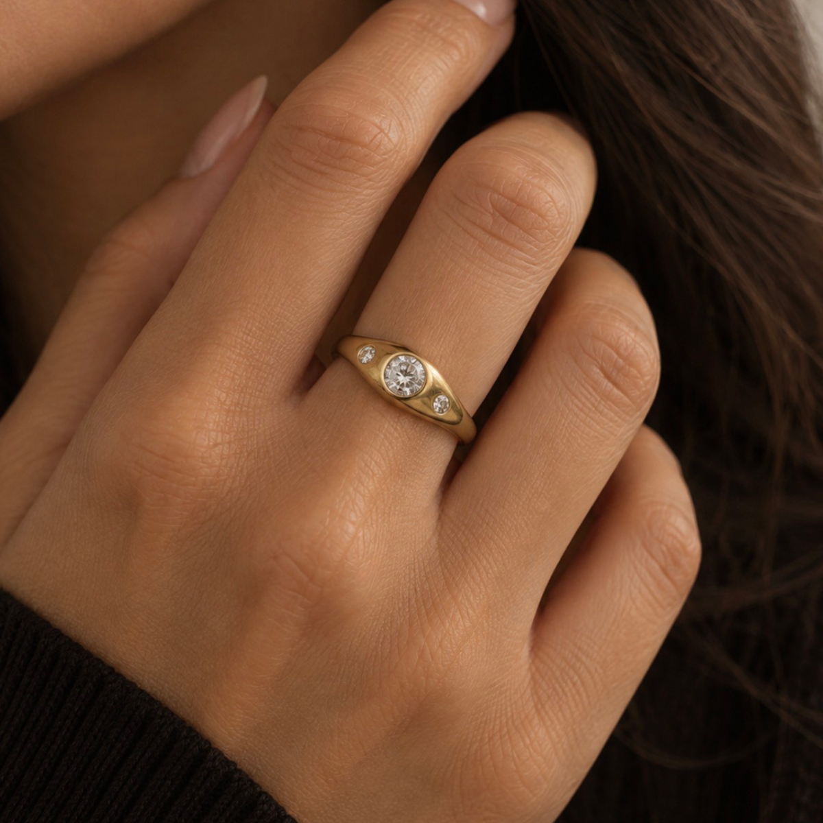 A close-up of a person's hand wearing a yellow gold ring with a round-cut center stone and two small side stones, set against dark hair and a dark ribbed sweater.