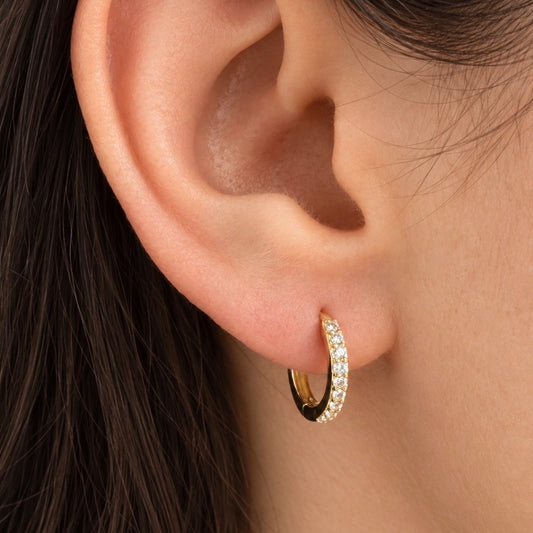 A close-up of a person's ear wearing a small gold huggie hoop earring set with clear pavé stones.