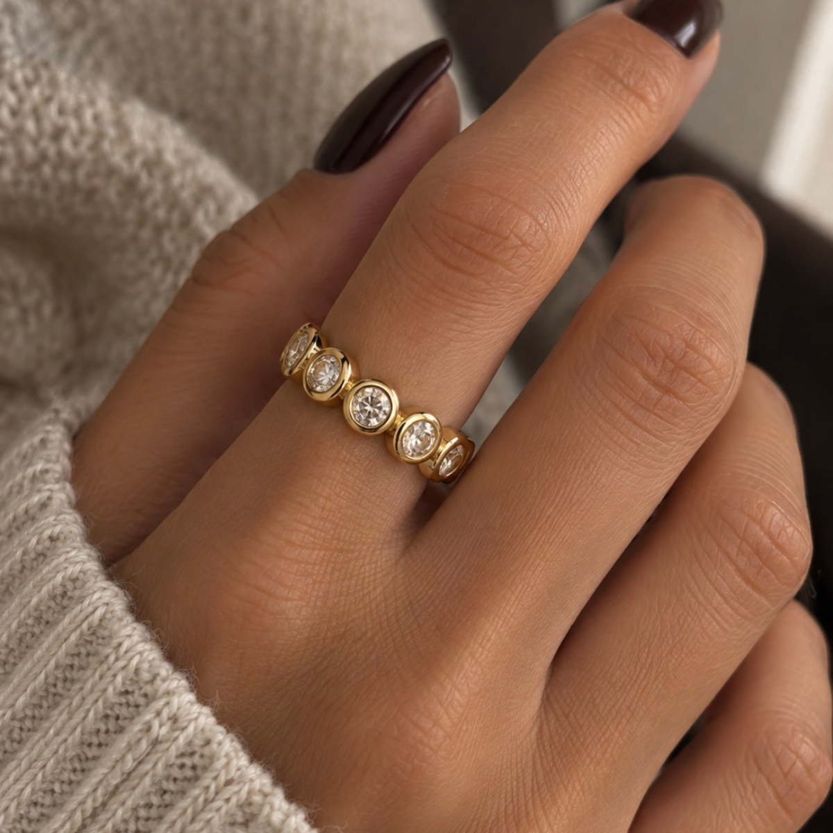A close-up of a hand wearing a gold eternity ring with round-cut clear gemstones, styled with a cream knit sweater.