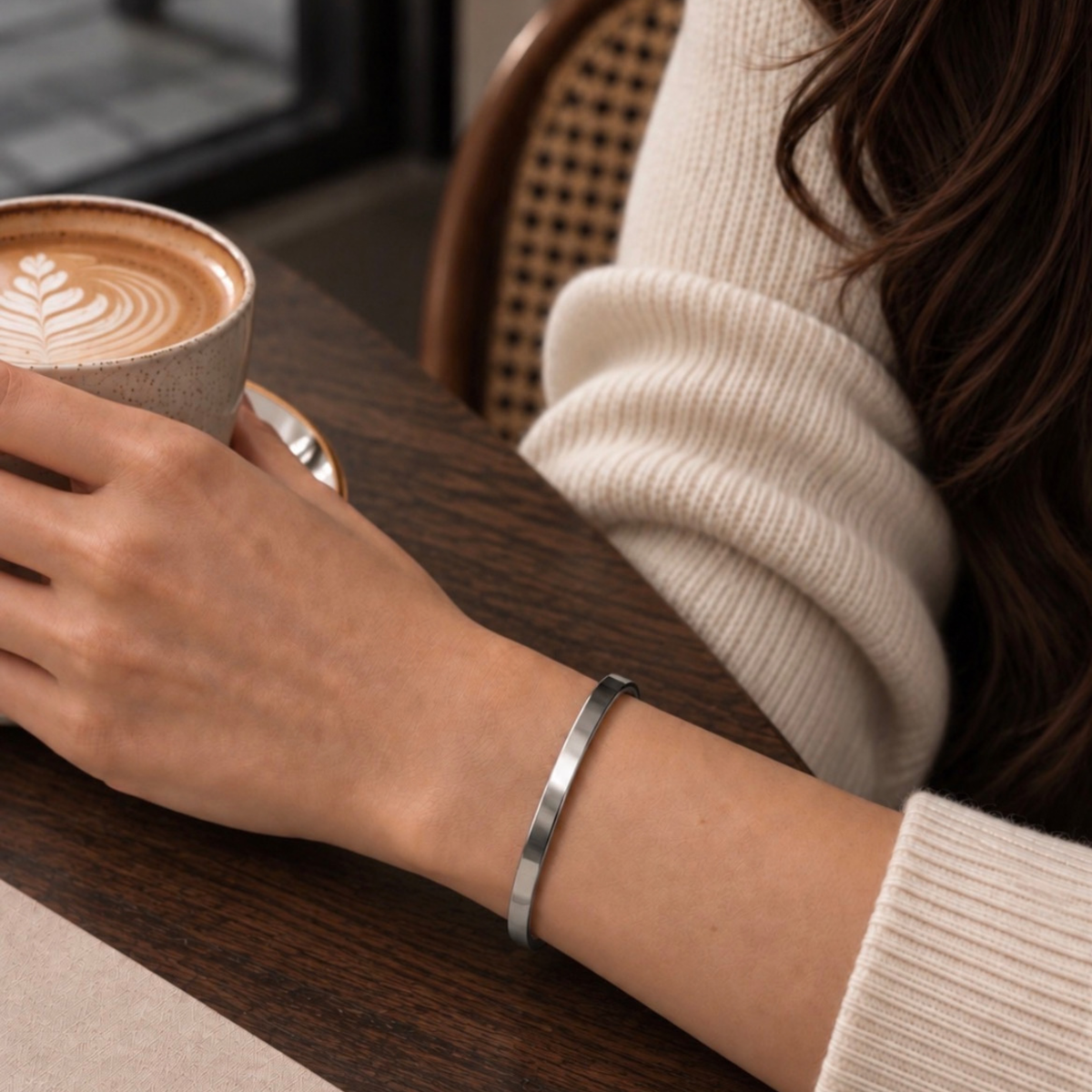 A close-up lifestyle shot of a woman wearing a slim, polished silver metal bangle bracelet on her wrist, seated at a wooden table in a cafe with a latte in a ceramic cup.