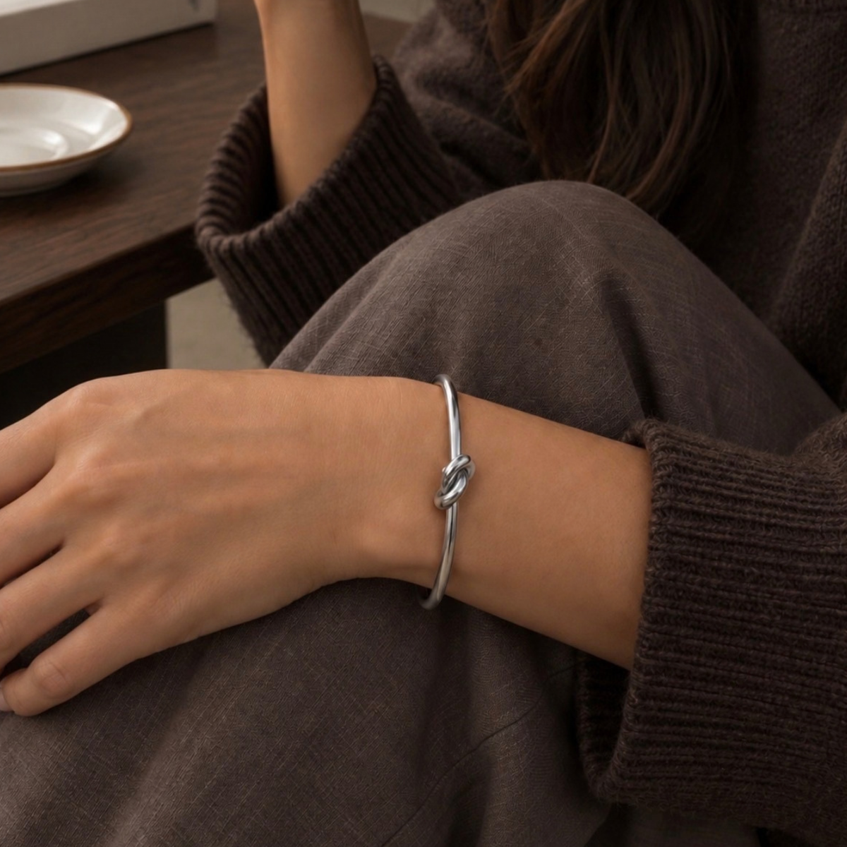 A close-up of a person's wrist wearing a thin silver bangle bracelet with a knotted detail, paired with a dark brown ribbed sweater and pants.